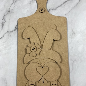 May include: A wooden Easter-themed cutting board with a bunny design. The bunny has large ears, a flower, and a heart-shaped nose. The board has a handle with a hole and a button detail. The bunny's overalls have button details and a heart.