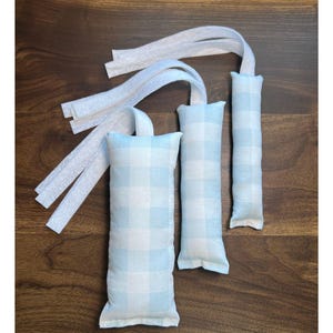 May include: Three rectangular, light blue and white checkered fabric sachets with white ribbon ties. The sachets are arranged on a dark wood surface. The fabric has a classic gingham pattern.