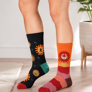 May include: Novelty socks featuring lion designs. One sock is black with orange and yellow lion faces, the other is orange with a lion graphic. The socks have red, green, and brown accents.