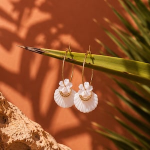 Peut inclure: Une paire de créoles dorées avec des pendentifs en forme d'éventail en coquillage blanc. Chaque pendentif est orné d'un petit croissant doré texturé sur le dessus, suspendu à un anneau fin en fil d'or passant par une petite fleur blanche.