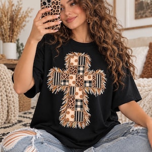 Puede incluir: Una camiseta negra de manga corta con un gran gráfico de cruz en la parte delantera. La cruz está compuesta por varios patrones de patchwork, incluyendo estampado de leopardo, cuadros en blanco y negro y lunares beige, con flecos.