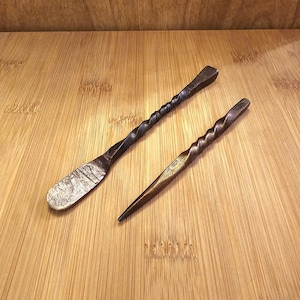 Peut inclure: Deux outils en m&eacute;tal forg&eacute; &agrave; la main. L'un est en forme de spatule avec une lame textur&eacute;e. L'autre est une al&egrave;ne pointue. Les deux ont des manches torsad&eacute;s et une finition sombre et vieillie. Ils reposent sur une surface en bois brun clair.