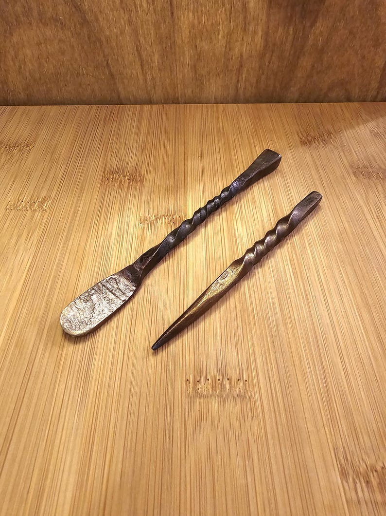 Peut inclure: Deux outils en m&eacute;tal forg&eacute; &agrave; la main. L'un est en forme de spatule avec une lame textur&eacute;e. L'autre est une al&egrave;ne pointue. Les deux ont des manches torsad&eacute;s et une finition sombre et vieillie. Ils reposent sur une surface en bois brun clair.