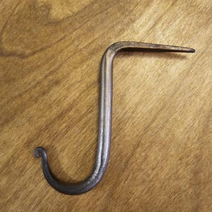 Könnte beinhalten: Handgeschmiedeter Metallhaken mit einem gebogenen, dekorativen Ende und einer geraden, spitzen Verlängerung. Der Haken ist silberfarben mit dunkleren Bereichen, wahrscheinlich vom Schmiedeprozess.