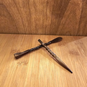 Peut inclure: Deux b&acirc;tonnets &agrave; cheveux en bois faits &agrave; la main avec une finition fonc&eacute;e. L'un a un design torsad&eacute;, l'autre une extr&eacute;mit&eacute; plate en forme de pagaie. Les accessoires pour cheveux sont pr&eacute;sent&eacute;s sur une surface en bois.