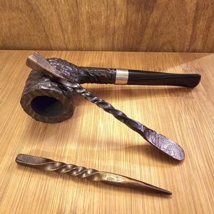 May include: A black tobacco pipe with a silver band, alongside two hand-forged metal tools. One tool features a twisted handle and a spoon-like end, the other is a poker with a twisted handle. The items are displayed on a wooden surface.