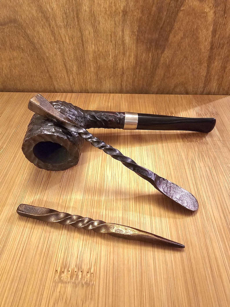 Peut inclure: Une pipe &agrave; tabac noire avec une bague argent&eacute;e, accompagn&eacute;e de deux outils en m&eacute;tal forg&eacute;s &agrave; la main. L'un a une poign&eacute;e torsad&eacute;e et une extr&eacute;mit&eacute; en forme de cuill&egrave;re, l'autre est un tisonnier &agrave; poign&eacute;e torsad&eacute;e. Les objets sont pr&eacute;sent&eacute;s sur une surface en bois.