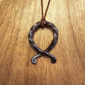 May include: Hand-forged metal pendant necklace with a twisted, looped design. The pendant is dark gray with a brown leather cord. The necklace is displayed against a wooden background.