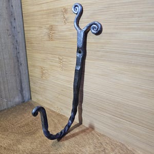 May include: Hand-forged iron hook with a dark, textured finish. The hook features a decorative swirl design at the top and a curved hook at the bottom. The metalwork is mounted on a light brown wooden surface.