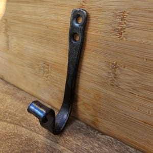 Könnte beinhalten: Handgeschmiedeter, dunkler Metallhaken mit geschwungenem Design. Der Haken hat zwei Befestigungslöcher oben und ein eingerolltes Ende. Der Haken wird vor einem Holzgrund gezeigt.
