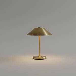 May include: A brass-colored table lamp with a conical shade and a slender stem. The lamp has a circular base and a warm, inviting glow emanating from the light source. The shade has a series of small perforations around the edge.