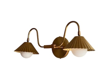 May include: A brass-toned wall sconce with two lights. Each light has a pleated shade and a white globe bulb. The fixture has a curved arm design and a circular base for mounting on the wall. The overall design is vintage-inspired.