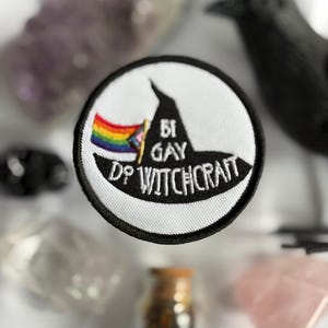 May include: A round, white patch with a black border featuring a witch's hat and the text "BI GAY DO WITCHCRAFT". A rainbow flag is incorporated into the design. The patch is surrounded by crystals, a black crow figurine, and small glass bottles.