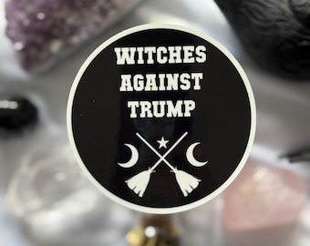 May include: A black and white sticker with the text "Witches Against Trump" and crossed broomsticks, moons, and a star design. The sticker is surrounded by crystals, a black bird figurine, and a small bottle.