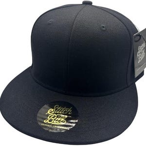 May include: A black baseball cap with a flat peak and a structured crown. The cap has a small button on top and a curved brim. A gold and black sticker on the brim reads "Stitch and Bill Classic Headwear". A black tag is attached.
