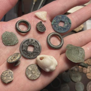 May include: A collection of antique coins and artifacts, including rings, a square-holed coin, and a white stone, are displayed in an open hand. The items vary in shape and size, with a mix of metallic and stone textures.
