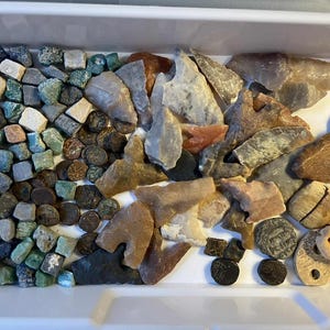 May include: A collection of various stones, including small, square-cut stones in shades of green, gray, and tan, alongside larger, irregularly shaped stones in brown, gray, and tan. Also included are small, round, dark-colored objects and carved, light-colored objects.