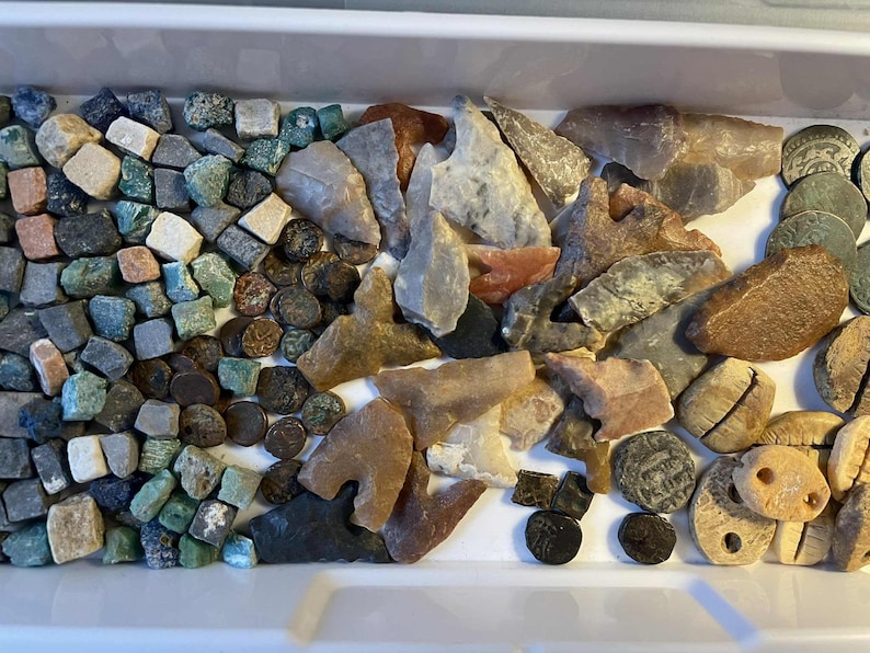 May include: A collection of various stones, including small, square-cut stones in shades of green, gray, and tan, alongside larger, irregularly shaped stones in brown, gray, and tan. Also included are small, round, dark-colored objects and carved, light-colored objects.