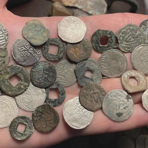May include: A collection of antique coins of various shapes and sizes, held in a hand. The coins are primarily silver and bronze, with visible inscriptions and designs. Some coins have square or circular holes in the center, and some show signs of wear and age.