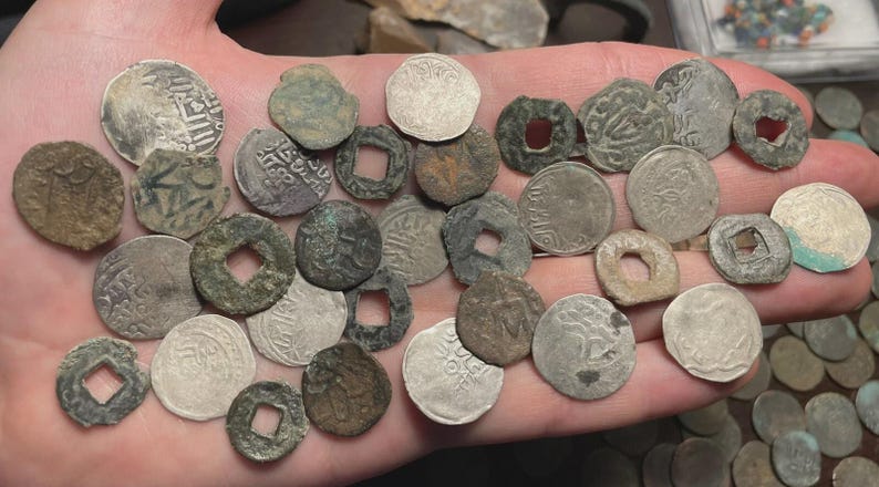May include: A collection of antique coins of various shapes and sizes, held in a hand. The coins are primarily silver and bronze, with visible inscriptions and designs. Some coins have square or circular holes in the center, and some show signs of wear and age.