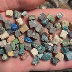May include: A collection of small, cube-shaped stones in various colors, including gray, green, blue, and beige. The stones are held in a hand, showcasing their diverse hues and textures. These appear to be small mosaic tiles or decorative stones.