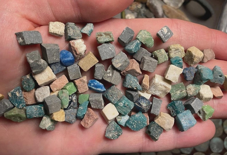 May include: A collection of small, cube-shaped stones in various colors, including gray, green, blue, and beige. The stones are held in a hand, showcasing their diverse hues and textures. These appear to be small mosaic tiles or decorative stones.