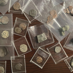 May include: A collection of ancient coins and artifacts, displayed in individual clear plastic sleeves. The coins vary in size, shape, and color, with visible details of their designs. Some artifacts are green and appear to be made of metal.