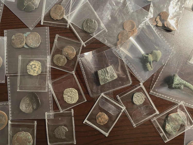 May include: A collection of ancient coins and artifacts, displayed in individual clear plastic sleeves. The coins vary in size, shape, and color, with visible details of their designs. Some artifacts are green and appear to be made of metal.