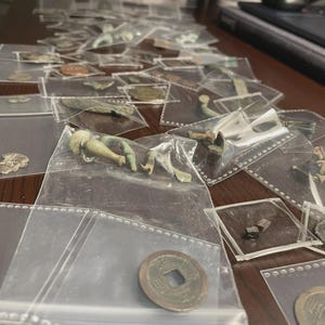 May include: A collection of ancient artifacts, including coins and small sculptures, are displayed in individual clear plastic sleeves. The items vary in size and shape, with a range of colors from bronze to green, suggesting age and historical significance.