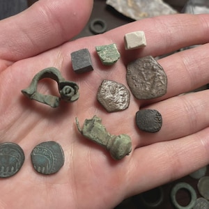 May include: A collection of ancient artifacts, including coins and fragments, in various shapes and sizes. The items are in shades of green, gray, and brown, with visible signs of age and wear. The objects are held in an open hand.