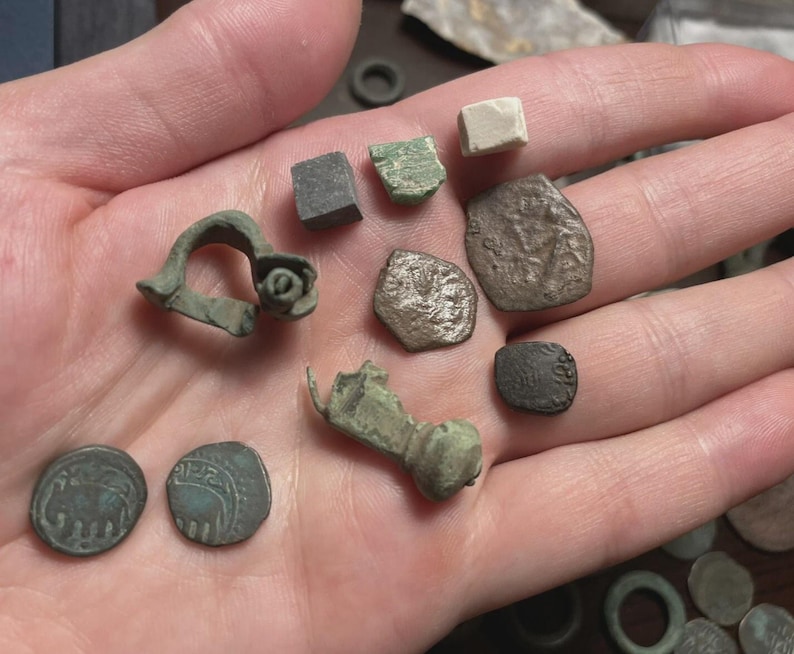 May include: A collection of ancient artifacts, including coins and fragments, in various shapes and sizes. The items are in shades of green, gray, and brown, with visible signs of age and wear. The objects are held in an open hand.