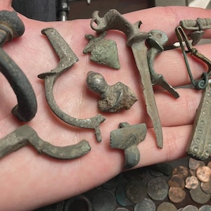 May include: A collection of antique metal artifacts, including clasps, pins, and decorative elements, in various shapes and sizes. The items have a weathered, aged appearance, with a mix of curved and angular designs. Some pieces show signs of wear and oxidation.
