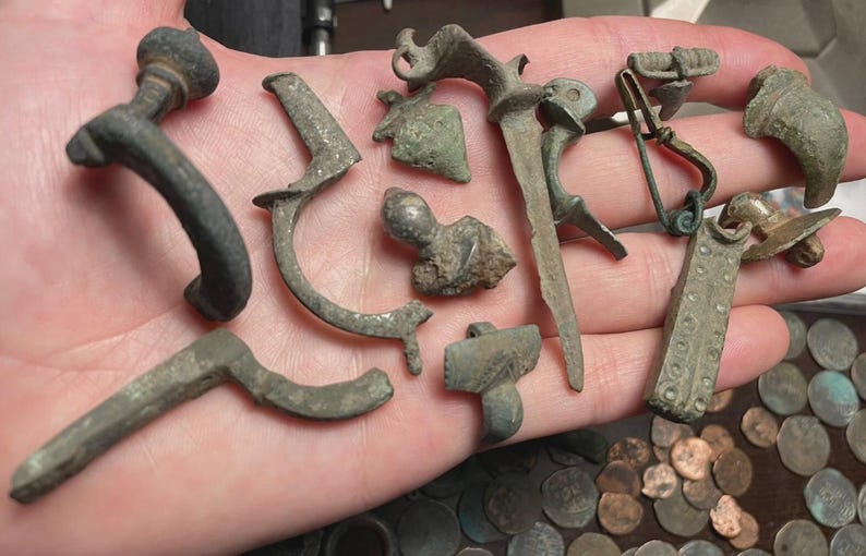 May include: A collection of antique metal artifacts, including clasps, pins, and decorative elements, in various shapes and sizes. The items have a weathered, aged appearance, with a mix of curved and angular designs. Some pieces show signs of wear and oxidation.