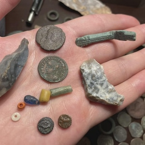 May include: A collection of ancient artifacts, including coins, beads, and stone tools, displayed in a hand. The coins are various sizes and shapes, with visible markings. The stone tools are gray and have sharp edges. The beads are colorful.