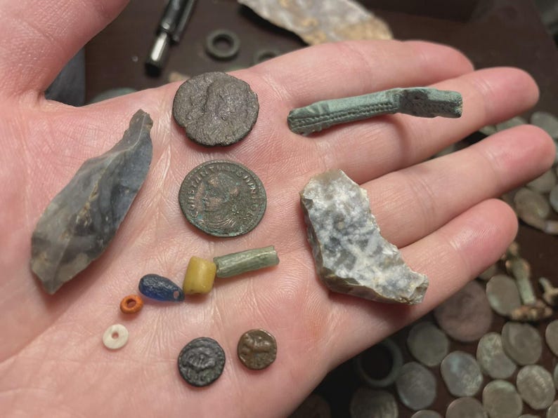 May include: A collection of ancient artifacts, including coins, beads, and stone tools, displayed in a hand. The coins are various sizes and shapes, with visible markings. The stone tools are gray and have sharp edges. The beads are colorful.