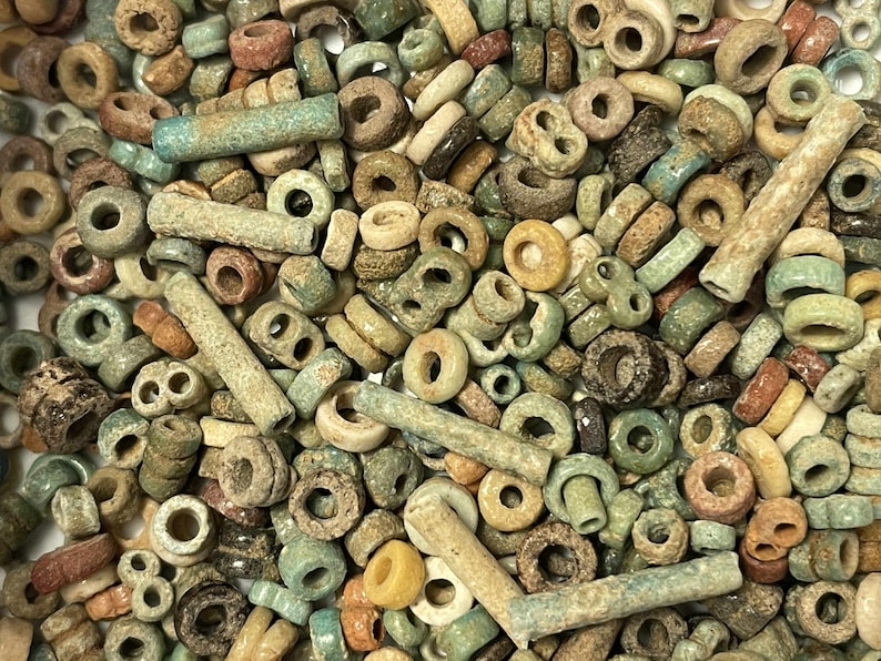 May include: A collection of small, antique beads in various shapes and sizes. The beads are primarily in shades of green, brown, and beige, with some showing signs of wear. The assortment includes round, tube, and other unique shapes, suggesting a vintage or handcrafted origin.