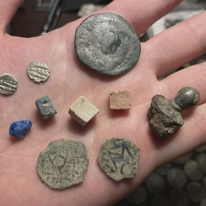 May include: A collection of ancient artifacts, including coins and small stone objects, are displayed in an open hand. The coins vary in size and shape, with some showing visible markings. A blue stone and a small cube are also present.
