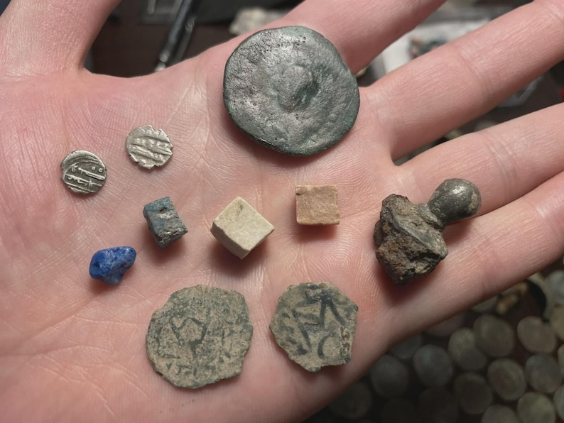 May include: A collection of ancient artifacts, including coins and small stone objects, are displayed in an open hand. The coins vary in size and shape, with some showing visible markings. A blue stone and a small cube are also present.
