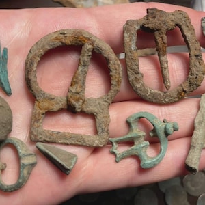 May include: A collection of antique metal artifacts, including buckles, arrowheads, and other historical items. The objects are in various states of preservation, with visible signs of age and wear. The color palette is primarily earthy tones.