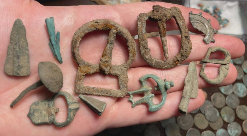 May include: A collection of antique metal artifacts, including buckles, arrowheads, and other historical items. The objects are in various states of preservation, with visible signs of age and wear. The color palette is primarily earthy tones.
