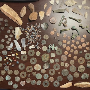 May include: A collection of ancient artifacts, including coins, beads, and stone tools, arranged on a dark wooden surface. The items vary in size, shape, and color, with some showing signs of wear and age. The assortment includes various coins and stone tools.