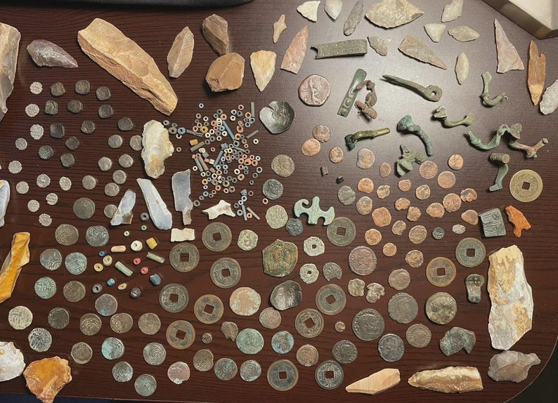 May include: A collection of ancient artifacts, including coins, beads, and stone tools, arranged on a dark wooden surface. The items vary in size, shape, and color, with some showing signs of wear and age. The assortment includes various coins and stone tools.