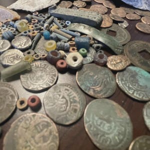 May include: A collection of antique coins, beads, and artifacts. The coins are silver and bronze, with intricate designs. Various colored beads and small stone tools are scattered among the coins. The items are arranged on a dark surface.