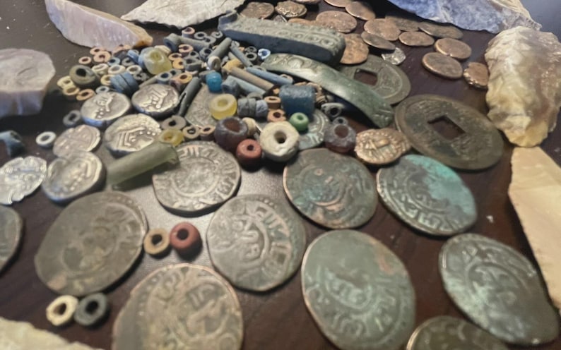 May include: A collection of antique coins, beads, and artifacts. The coins are silver and bronze, with intricate designs. Various colored beads and small stone tools are scattered among the coins. The items are arranged on a dark surface.