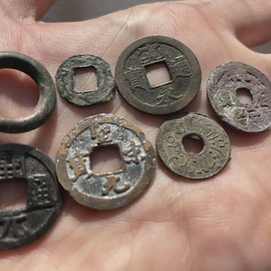 May include: Collection of antique Chinese coins and a ring, displayed on a hand. The coins are various sizes and shapes, with square holes in the center. The ring is dark gray. The coins show signs of age and wear.