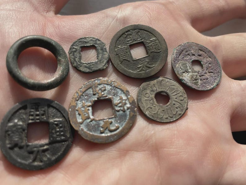 May include: Collection of antique Chinese coins and a ring, displayed on a hand. The coins are various sizes and shapes, with square holes in the center. The ring is dark gray. The coins show signs of age and wear.