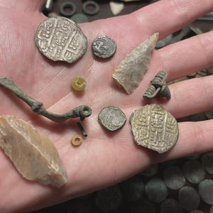 May include: A collection of ancient artifacts, including coins, a stone arrowhead, and a metal brooch, displayed in a hand. The coins have intricate designs, and the arrowhead is a light beige color. The brooch is a dark, aged metal.