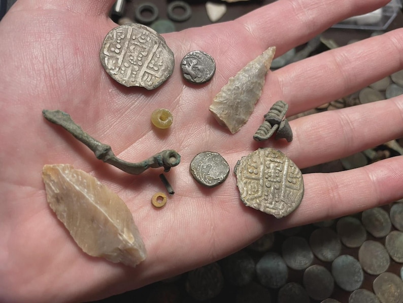 May include: A collection of ancient artifacts, including coins, a stone arrowhead, and a metal brooch, displayed in a hand. The coins have intricate designs, and the arrowhead is a light beige color. The brooch is a dark, aged metal.