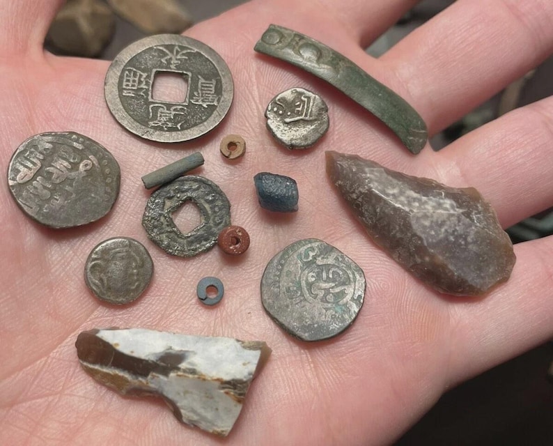 May include: A collection of antique coins and artifacts, including a Chinese coin with square hole, various round coins, a small blue stone, and a piece of flint. The items are in various shapes and sizes, with visible wear and tear.
