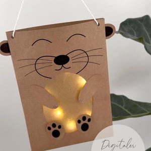 May include: A brown paper craft hanging decoration featuring a cartoon animal face with whiskers, ears, and a glowing yellow circular belly. The item includes a white string for suspension and displays the text Digitaler DOWNLOAD with a printer icon.
